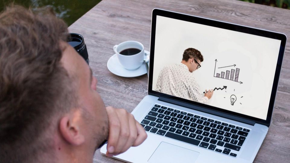 A man looking at a laptop screen that shows another man drawing on a whiteboard.